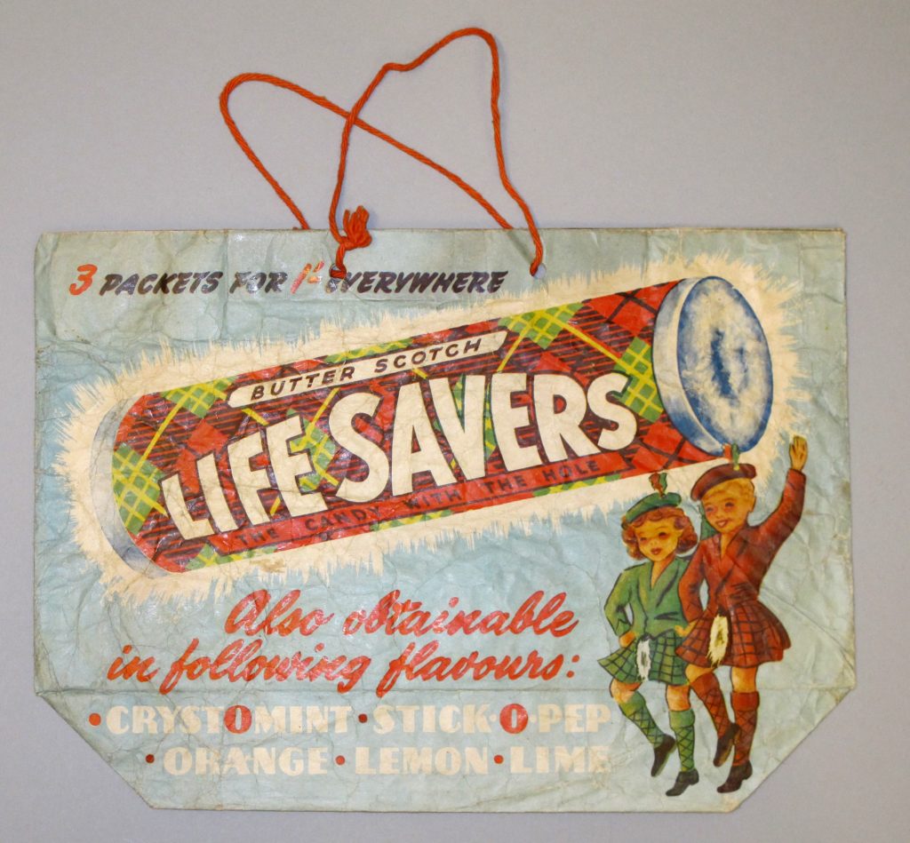 A paper bag with cotton cord handles featuring a coloured picture of a large roll of butterscotch lifesavers and two Scottish girls dancing in kilts.